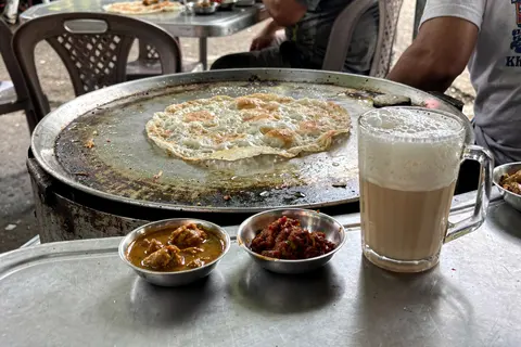 AI illustration: Freshly griddled roti canai with curry dips and a frothy glass of teh tarik at a Malaysian mamak stall in natural daylight.