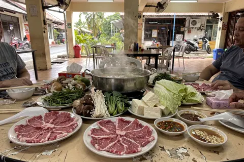 AI illustration: Steaming shared hotpot with sliced meat and fresh vegetables on a Malaysian restaurant table.