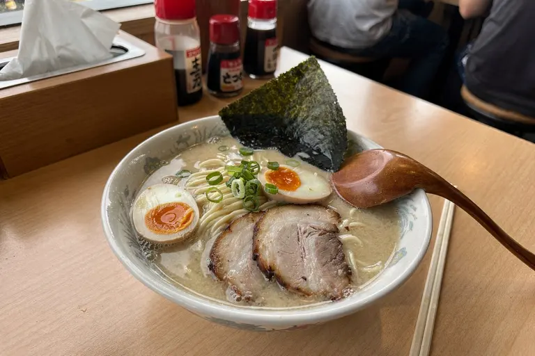 AI illustration: A close-up bowl of creamy tonkotsu ramen with chashu, ajitama egg, spring onions and nori on a wooden counter