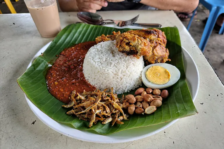 AI illustration: A plate of nasi lemak with coconut rice, sambal, anchovies, peanuts, boiled egg and fried chicken in natural light