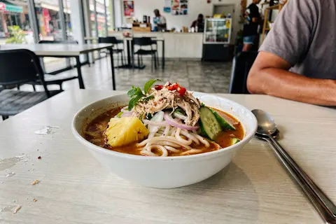 AI illustration: A bowl of vegan asam laksa with rice noodles, pineapple and herbs on a cafe table in George Town