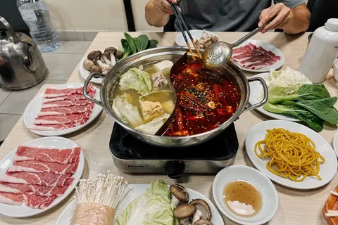 AI illustration: A divided hotpot of clear and spicy broth on a table with plates of thinly sliced beef, vegetables and dipping sauces
