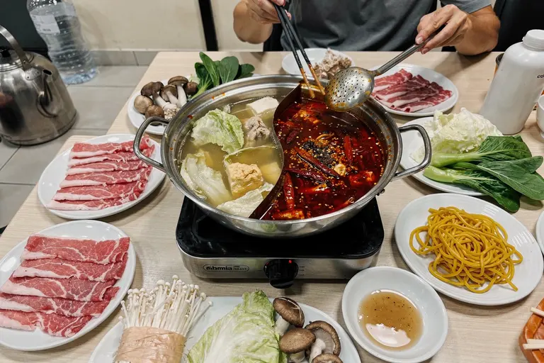 AI illustration: A divided hotpot of clear and spicy broth on a table with plates of thinly sliced beef, vegetables and dipping sauces
