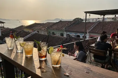 AI illustration: Golden-hour view from a Penang rooftop bar over George Town's heritage rooflines toward the glowing sea strait